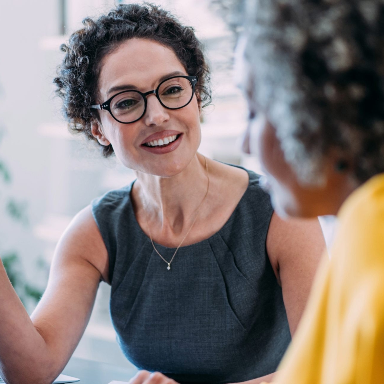 A white woman with short dark hair, weatring a grey top and glasses is holding a pen and sitting at a laptop is talking to a woman who is side on to the camera and wearing a yello top